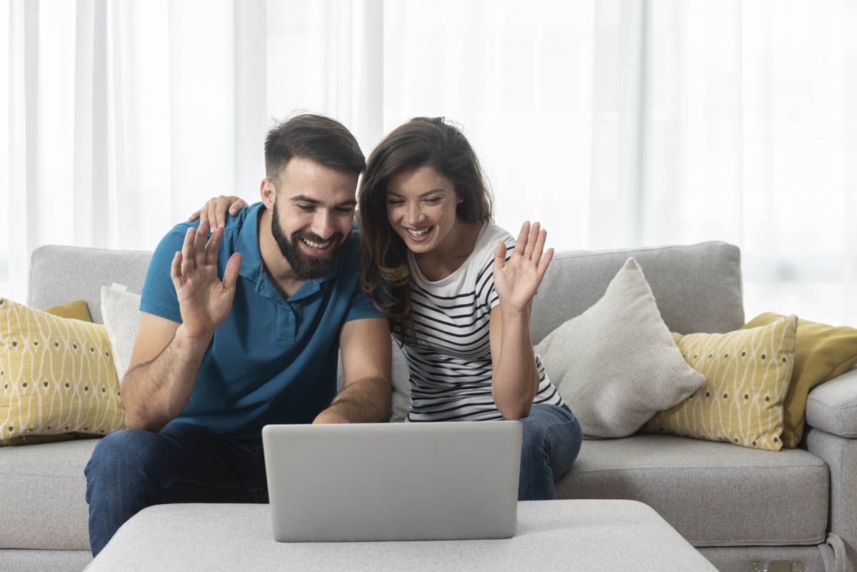 Young couple sitting in their living room with laptop waving to