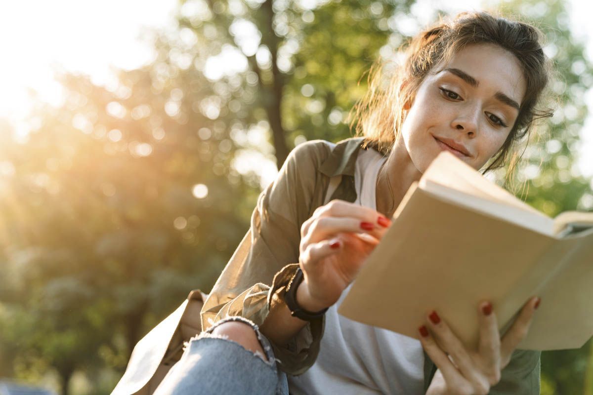 Image of young woman wearing casual clothes smiling and reading book while walking in green park